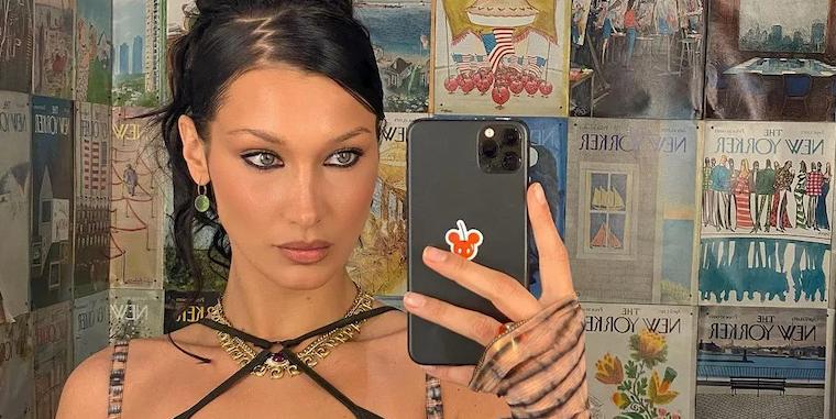 Modelo Bella Hadid aparece em frente a um espelho, enquanto segura um celular preto. Ela usa roupas, acessórios e penteado no estilo anos 2000.