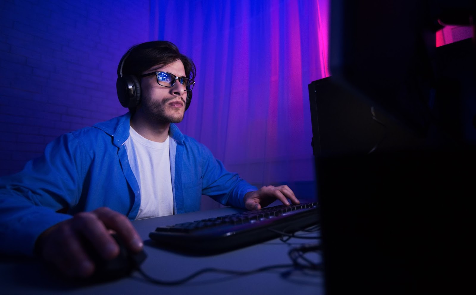 A man wearing blue light glasses plays video games in a dark room
