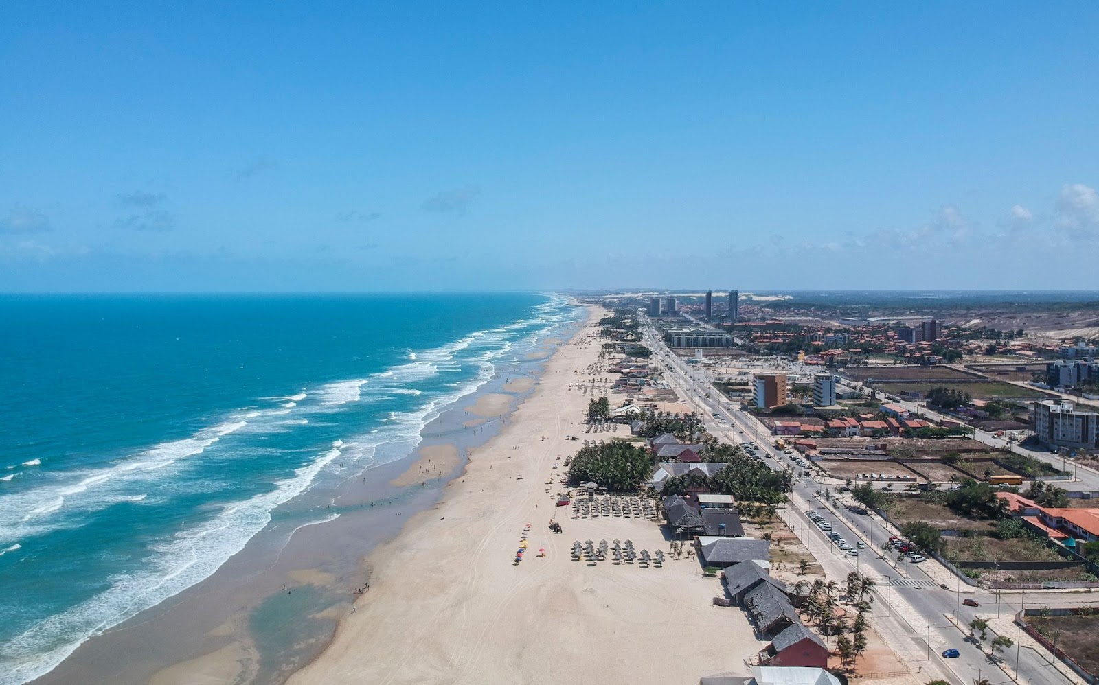 Praia do Futuro, em Fortaleza,  vista de cima. A extensa faixa de areia branca é ocupada por quiosques e guarda-sóis, mas tem muitas áreas vazias. O mar e o céu têm uma coloração azul vibrante.