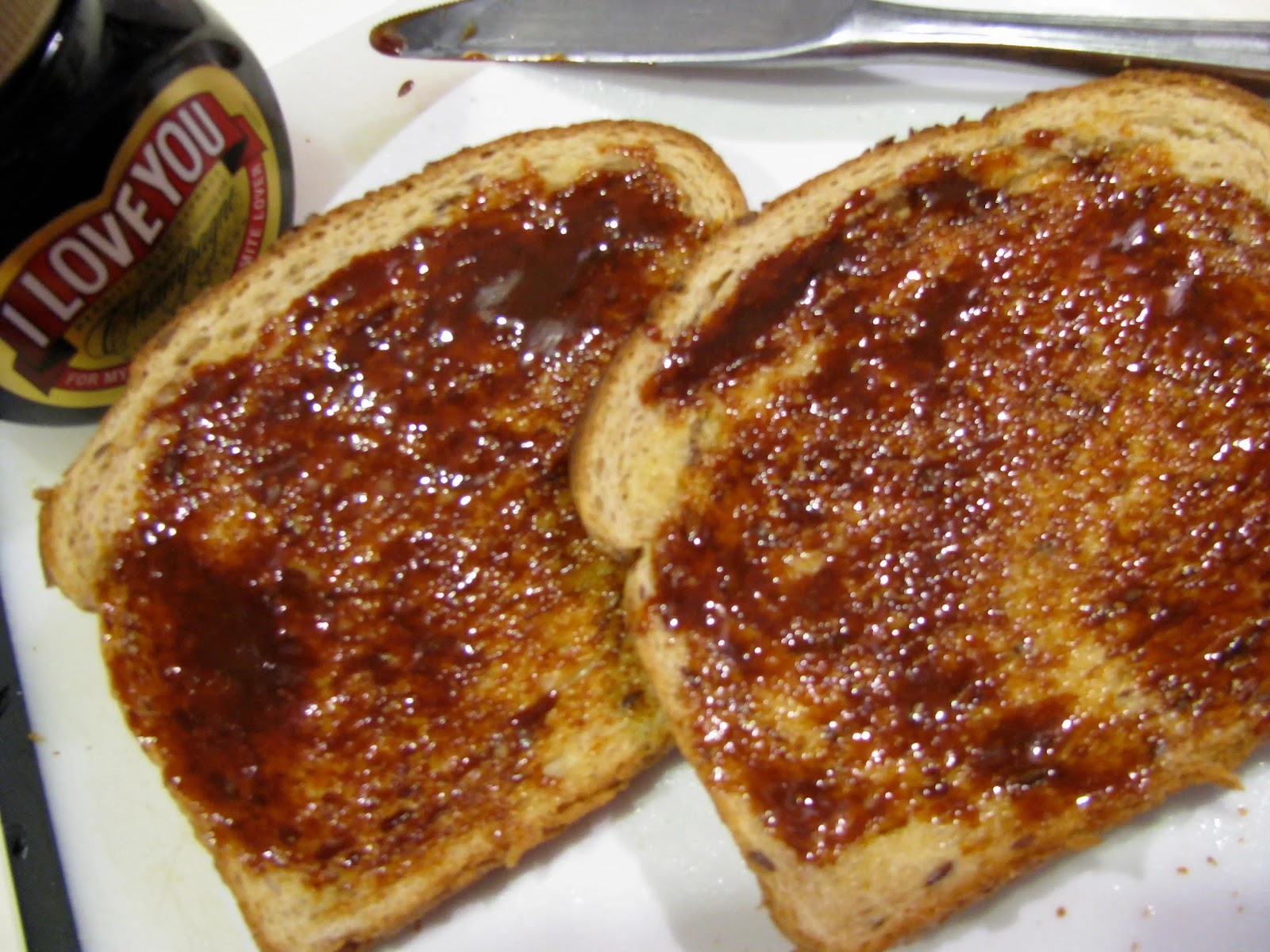 A thick slice of golden brown sourdough toast, generously spread with melting butter and a thin layer of dark brown Marmite, served on a white ceramic plate with a silver butter knife resting beside it.