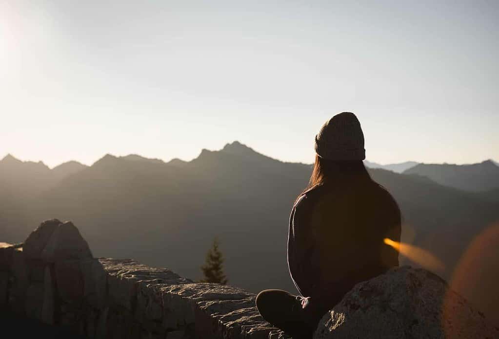 A person sits on a rock wall, facing away from the camera, overlooking a mountain range. The individual is wearing a beanie and appears to be deep in contemplation as the sun rises or sets, casting a soft, warm light over the mountains and creating a serene atmosphere. The sky is clear, and the distant peaks are bathed in gentle light, adding to the tranquil scene.