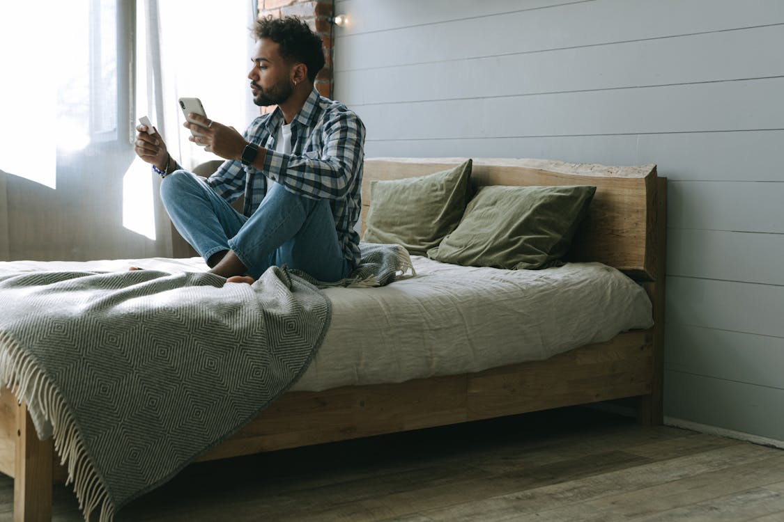 Free A Man in Checkered Long Sleeves Sitting on Bed While Holding Cellphone Stock Photo