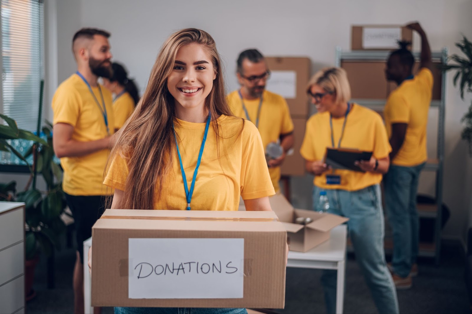 A group of volunteers in yellow shirts packing food while a smiling young woman holds a donation box.
