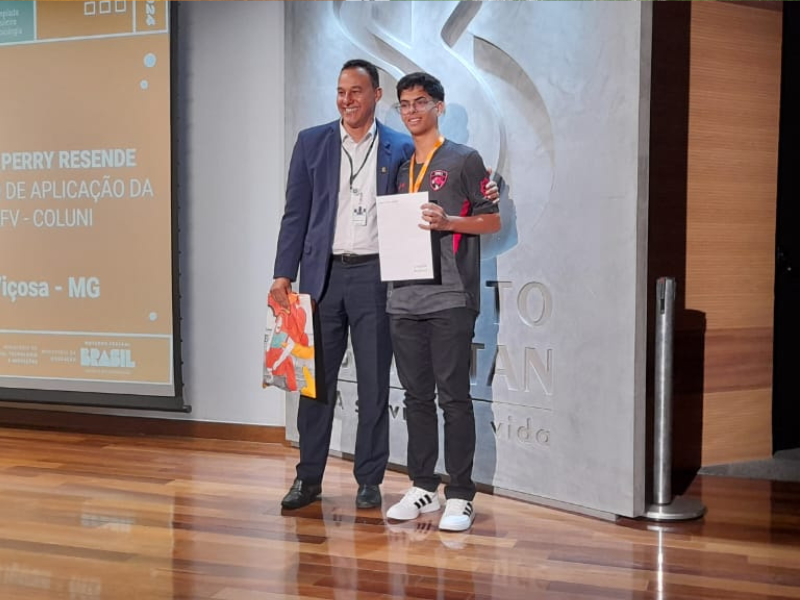 Estudante do Coluni representa Viçosa internacionalmente.

Imagem de Pedro Perry recebendo um certificado na sede do Instituto Butantan

A imagem mostra dois homens em um palco durante um evento de premiação. À esquerda, um homem de terno escuro, camisa branca e gravata sorri enquanto segura uma sacola colorida. À direita, um jovem de óculos e cabelo curto veste uma camiseta cinza escuro com detalhes vermelhos e calça preta, segurando um certificado. Eles estão lado a lado, com o braço do homem mais velho em volta do jovem. Ao fundo, um painel com a logomarca do evento e uma parede com o logo do Instituto Federal. No canto inferior esquerdo do painel de fundo, está escrito: "Perry Resende, Colégio de Aplicação da UFV-COLUNI, Viçosa - MG".