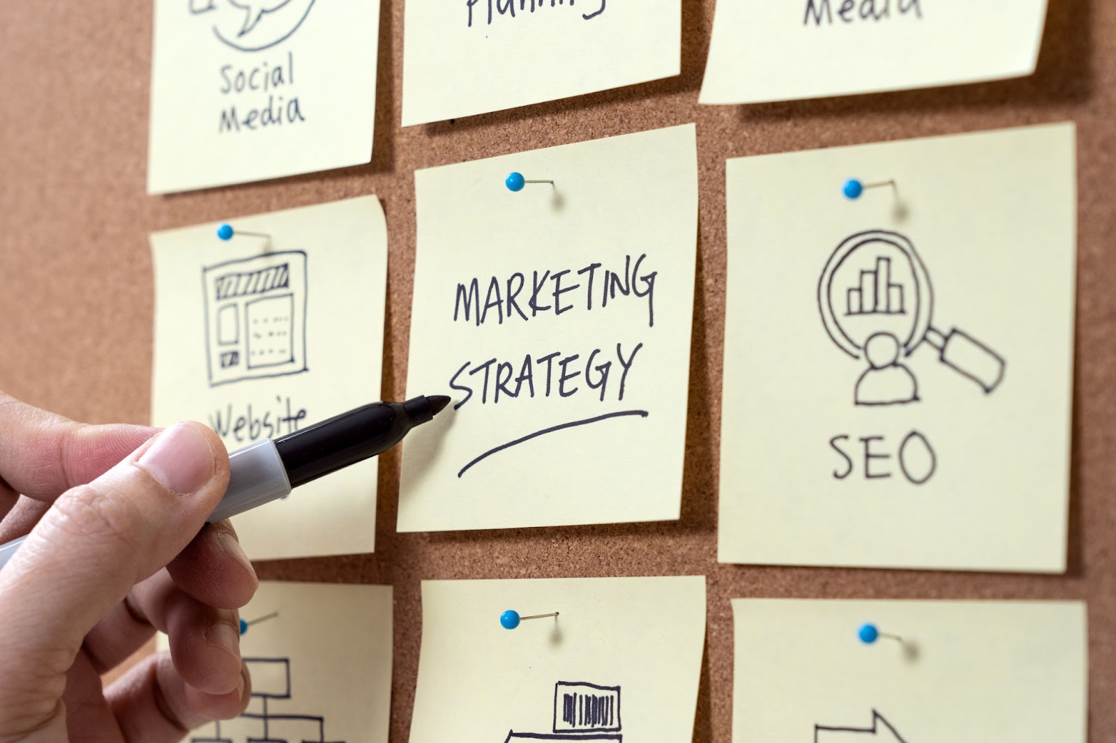 A male hand holds a marker, highlighting "marketing strategy" in the center of steps written on sticky yellow notes and piecing them together on a corkboard.