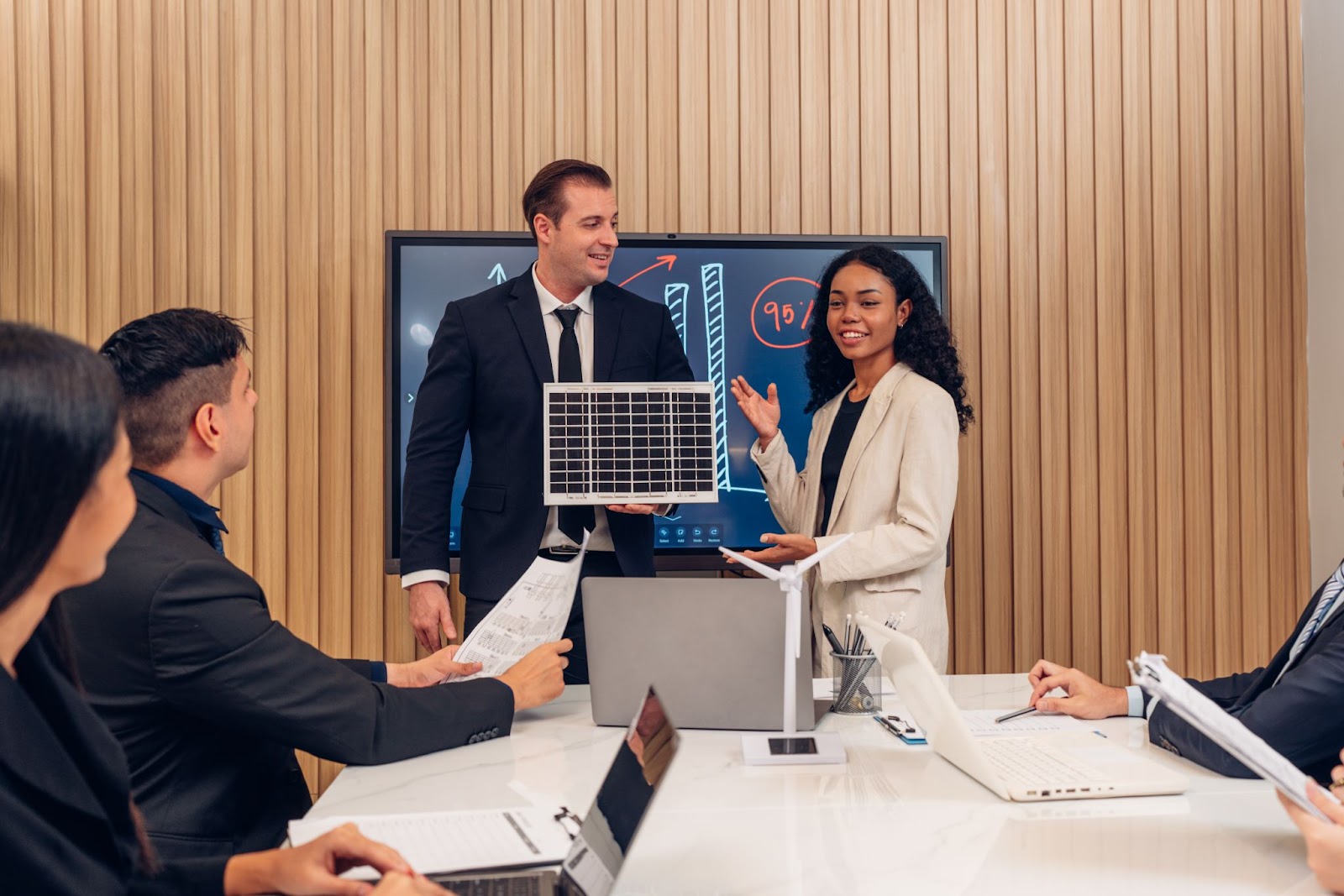 Two business people present solar cell panel technology to a panel with laptops on the table, ensuring the technology meets compliance. 