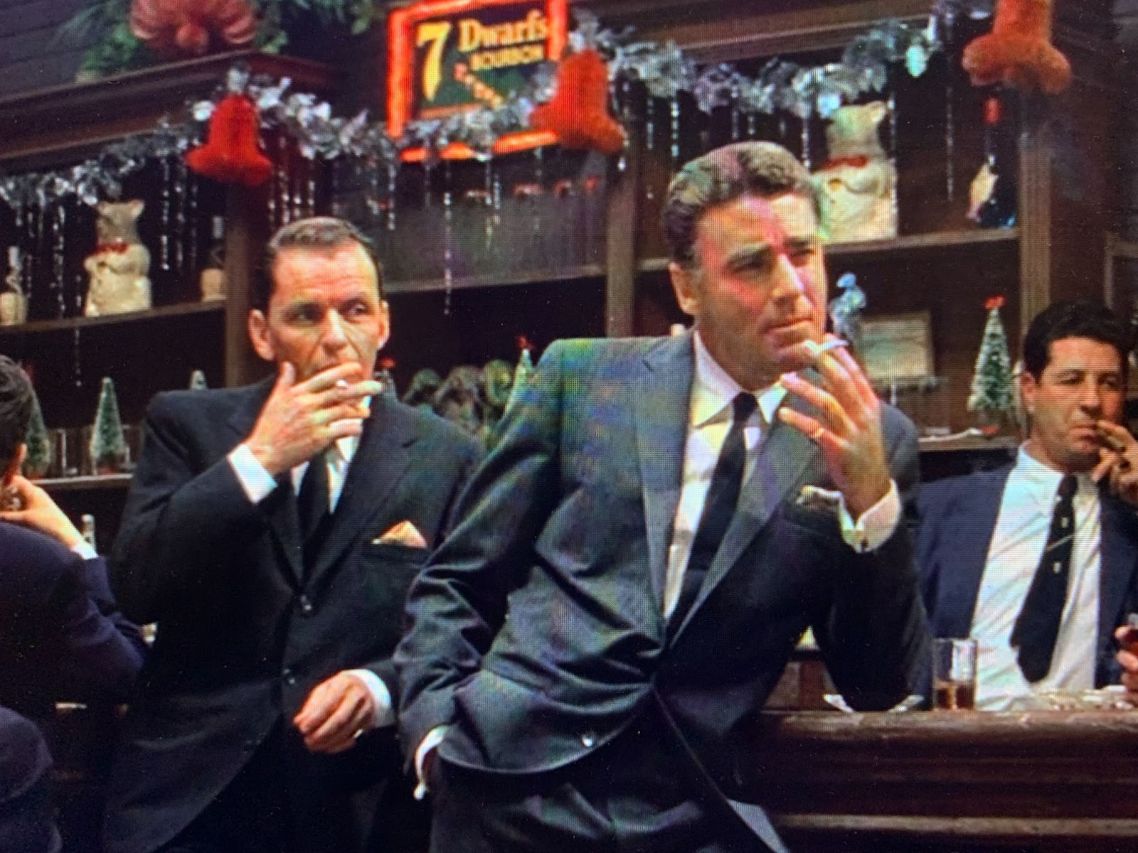 Peter Lawford, in a dark suit with a thin black tie, leans with his right hand in his hip pants pocket, his left elbow on the bar, and a cigarette held at chin level between the index and middle of the otherwise splayed and slightly curved fingers of his left hand. He has a pinky ring. Just behind him, to the left, Frank Sinatra in a darker suit, three-buttoned, with a peach-colored pocket square, white shirt, and dark tie, leans his left elbow on the bar and takes a drag on a cigarette tucked between the fingers of his splayed right hand. Both are looking off to their left. The smoking gestures help highlight both of their well-shot cuffs, Lawford's being French with a visible cufflink. Behind them the bar is hung with silver tinsel and red crepe-paper bells over a sign that reads "7 Dwarfs Bourbon." The bartender, at the right of the frame in an unbuttoned suit jacket, is facing the same way and has a cigarette raised to his mouth in his left hand.  