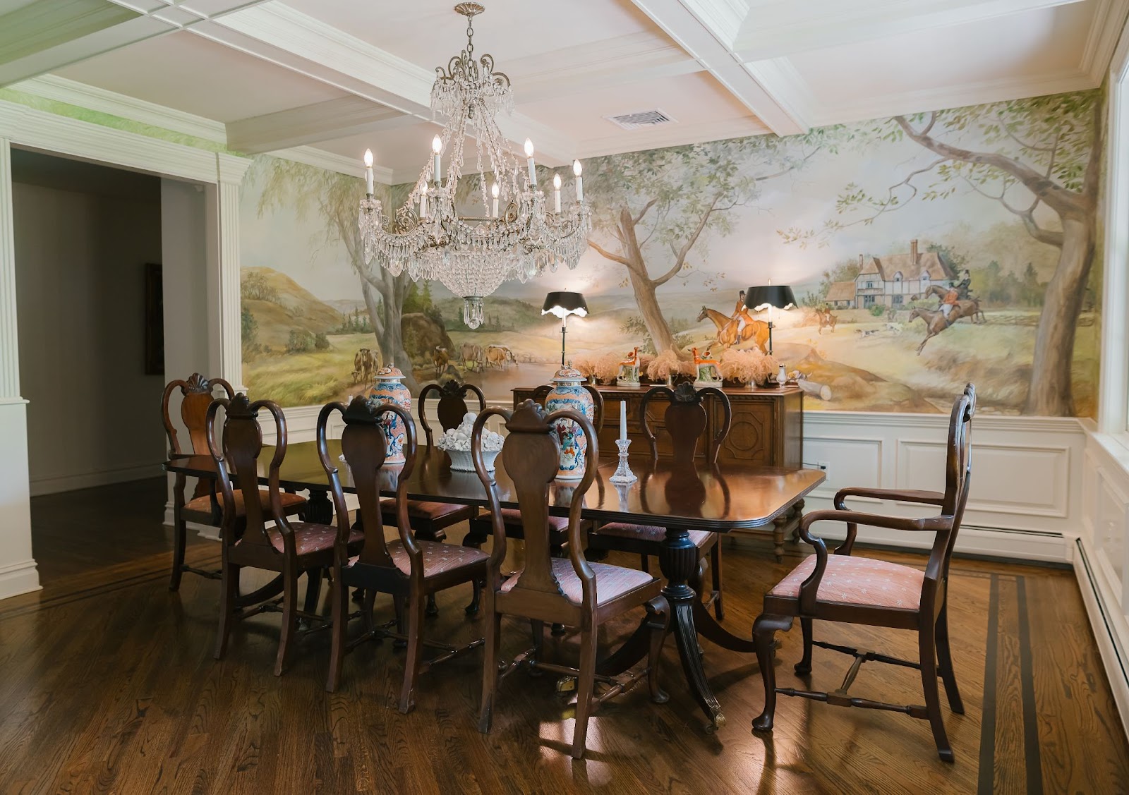 A dining room features coffered ceiling and pastoral mural wallpaper with a long mahogany dining  table surrounded by matching chairs and an ornate crystal chandelier.