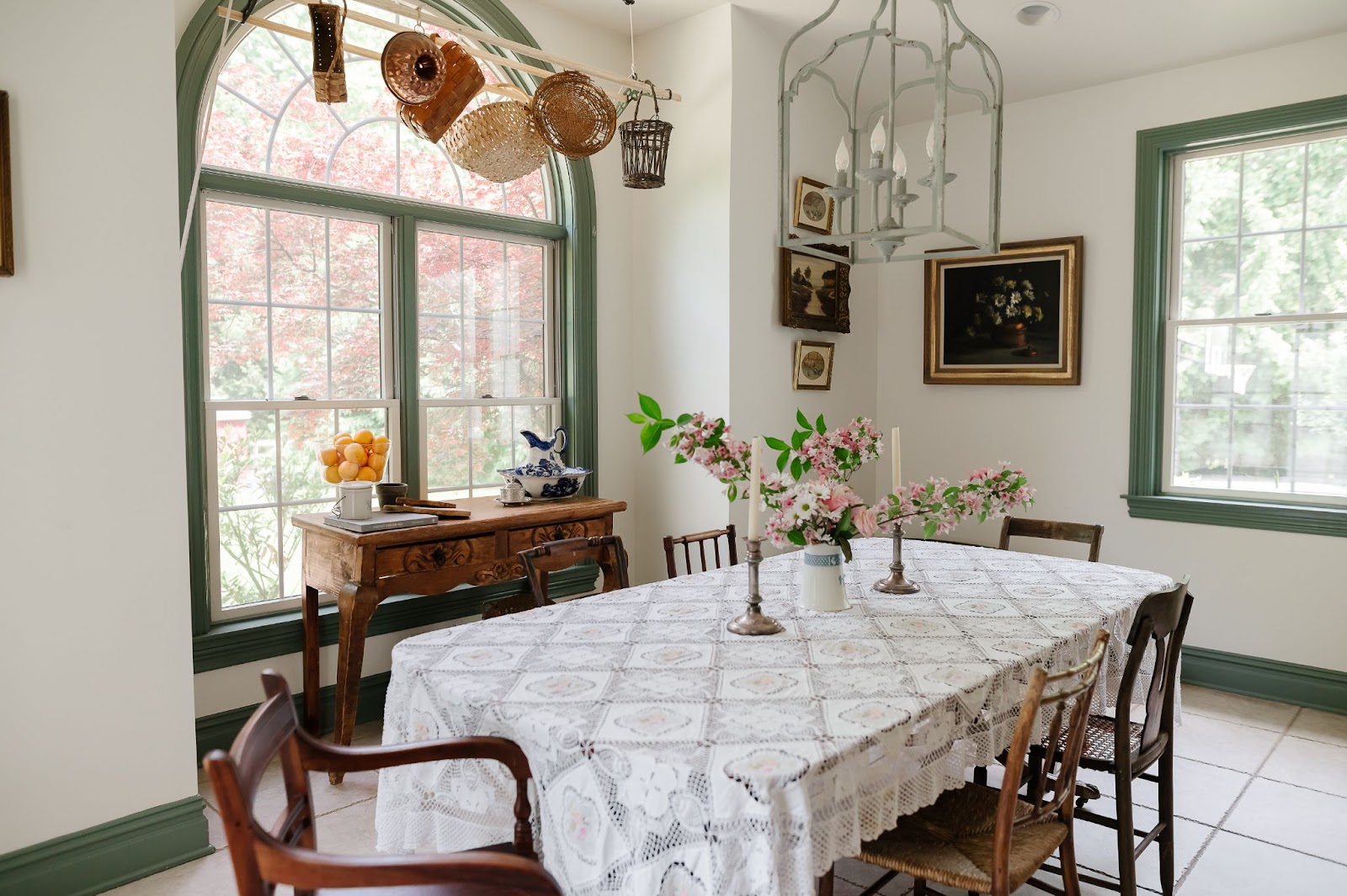 A light and airy dining area featuring white walls and green trim, a table covered in a lace tablecloth, floral centerpieces and candlesticks along with works of art on the walls.