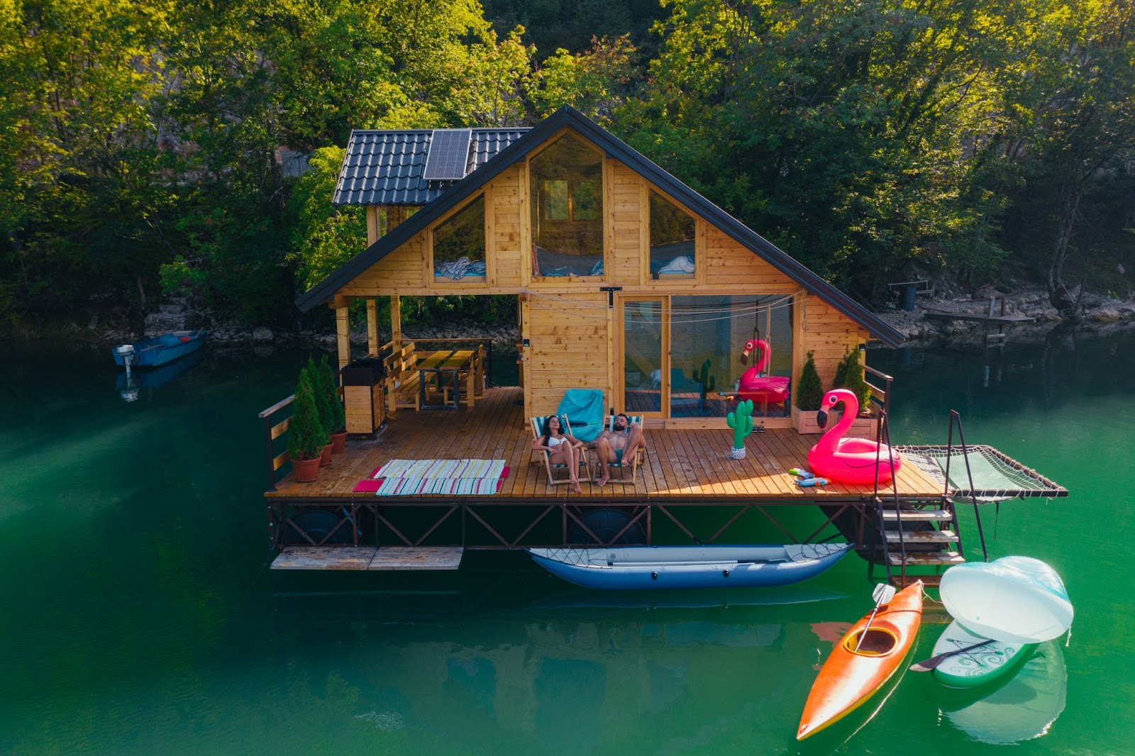 A floating house with a deck overlooking a pond, a small boat, and duck-shaped inflatable floaters.