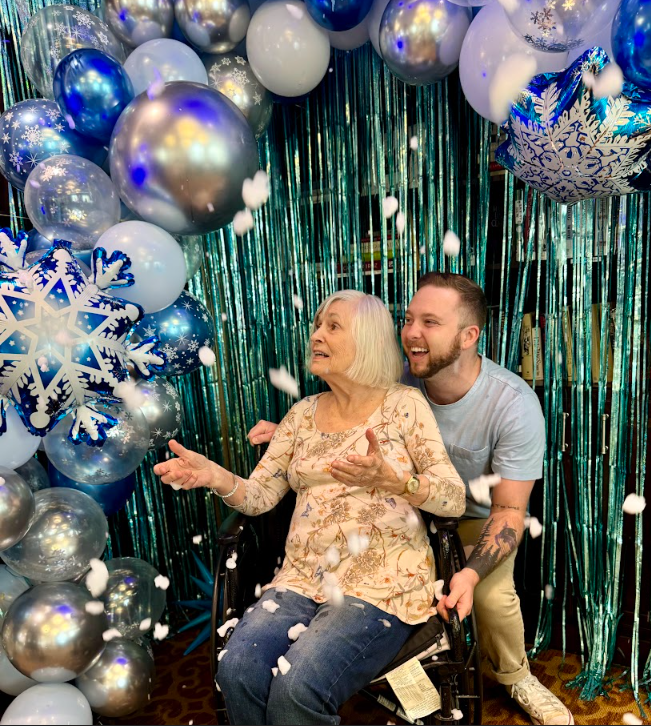 An elderly woman in a wheelchair smiling as fake snow is thrown in the air.