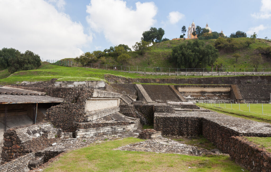 Ancient stone ruins of Cholula, Mexico, with grassy hills and a church atop a hill in the background under a partly cloudy sky.