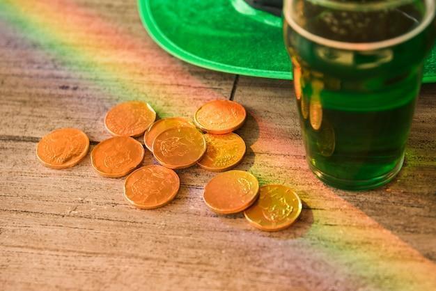 Glass of drink near heap of coins and Saint Patricks hat at table
