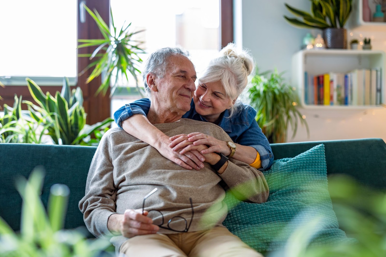 An older adult woman embraces her husband who sits on a couch surrounded by plants. They smile at each other.