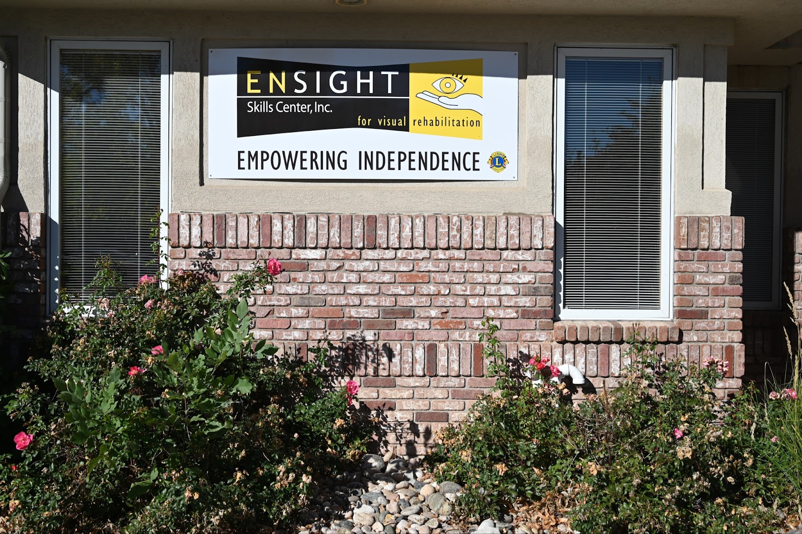 The image shows the exterior of a building with a sign that reads "ENSIGHT Skills Center, Inc. for visual rehabilitation" and the slogan "EMPOWERING INDEPENDENCE." The sign features a yellow and black color scheme, with an icon of an eye and a hand. The building is made of brick and has windows with closed blinds on either side of the sign.