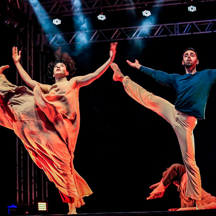 Dois dançarinos em um palco iluminado, fazendo movimentos graciosos e expressivos, com trajes fluídos