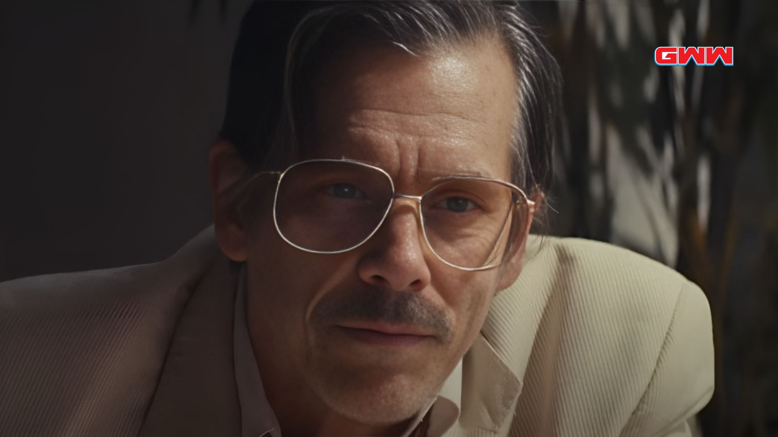 Kevin Bacon with glasses and mustache, wearing a beige suit.
