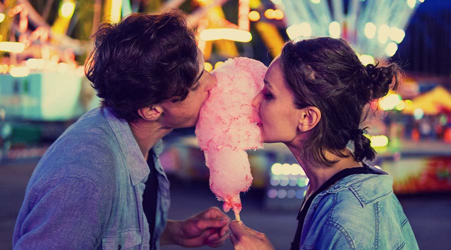 Brunette man and woman holding a stick of cotton candy between them and biting down on it from each side.
