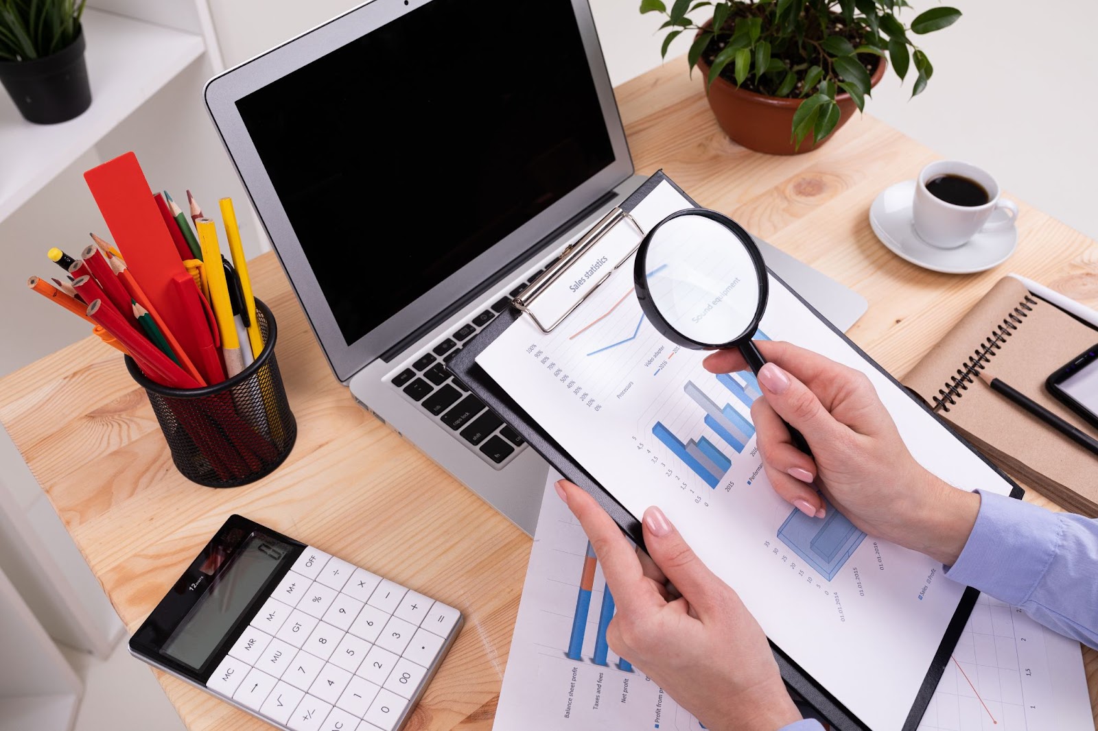 A compliance auditor reviews charts with a magnifying glass, working on a laptop at a desk filled with a calculator, pens, pencils, and cards.