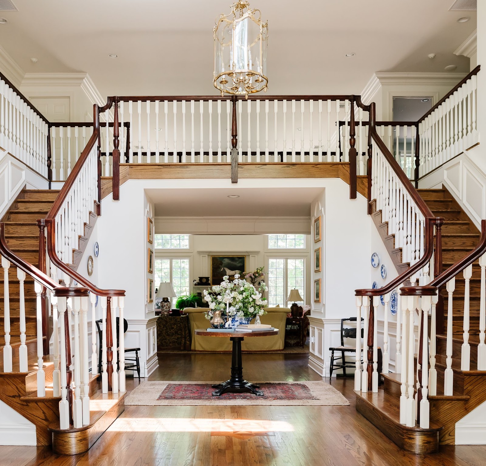 A grand foyer featuring a curved  double staircase, large chandelier, and a pedestal table with a large flower arrangement. A nicely decorated living room is in the background.