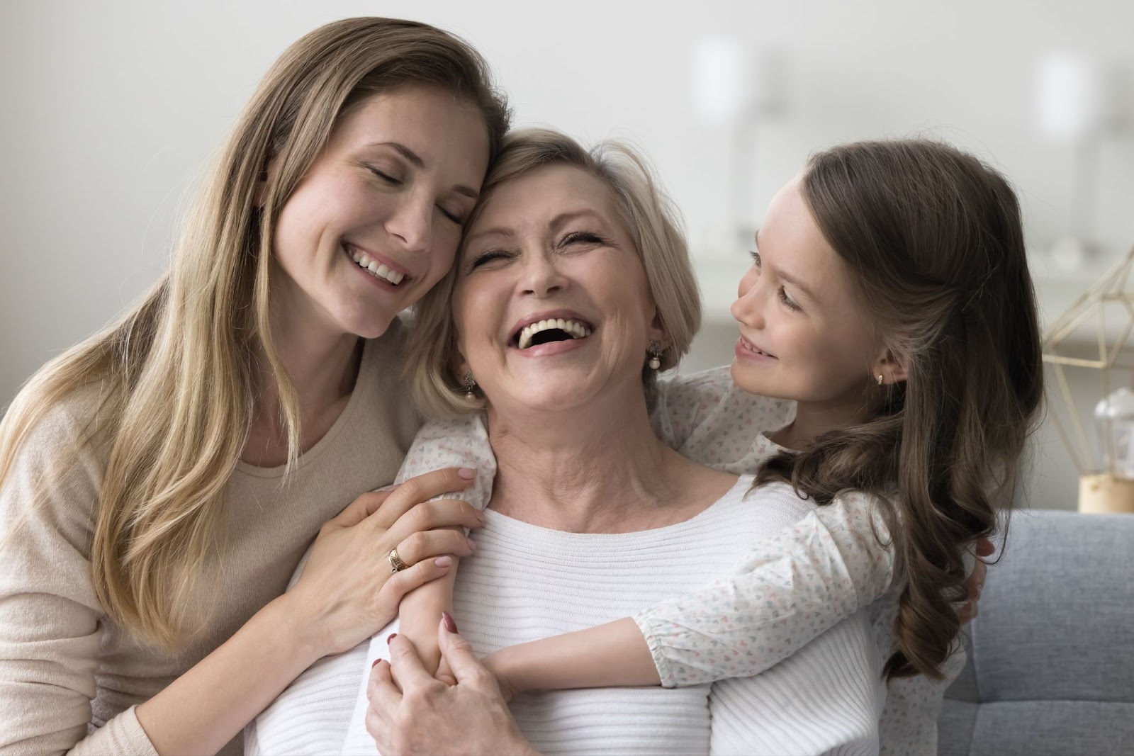 A woman and her daughter laugh and hug their happy grandma as the three sit on a couch