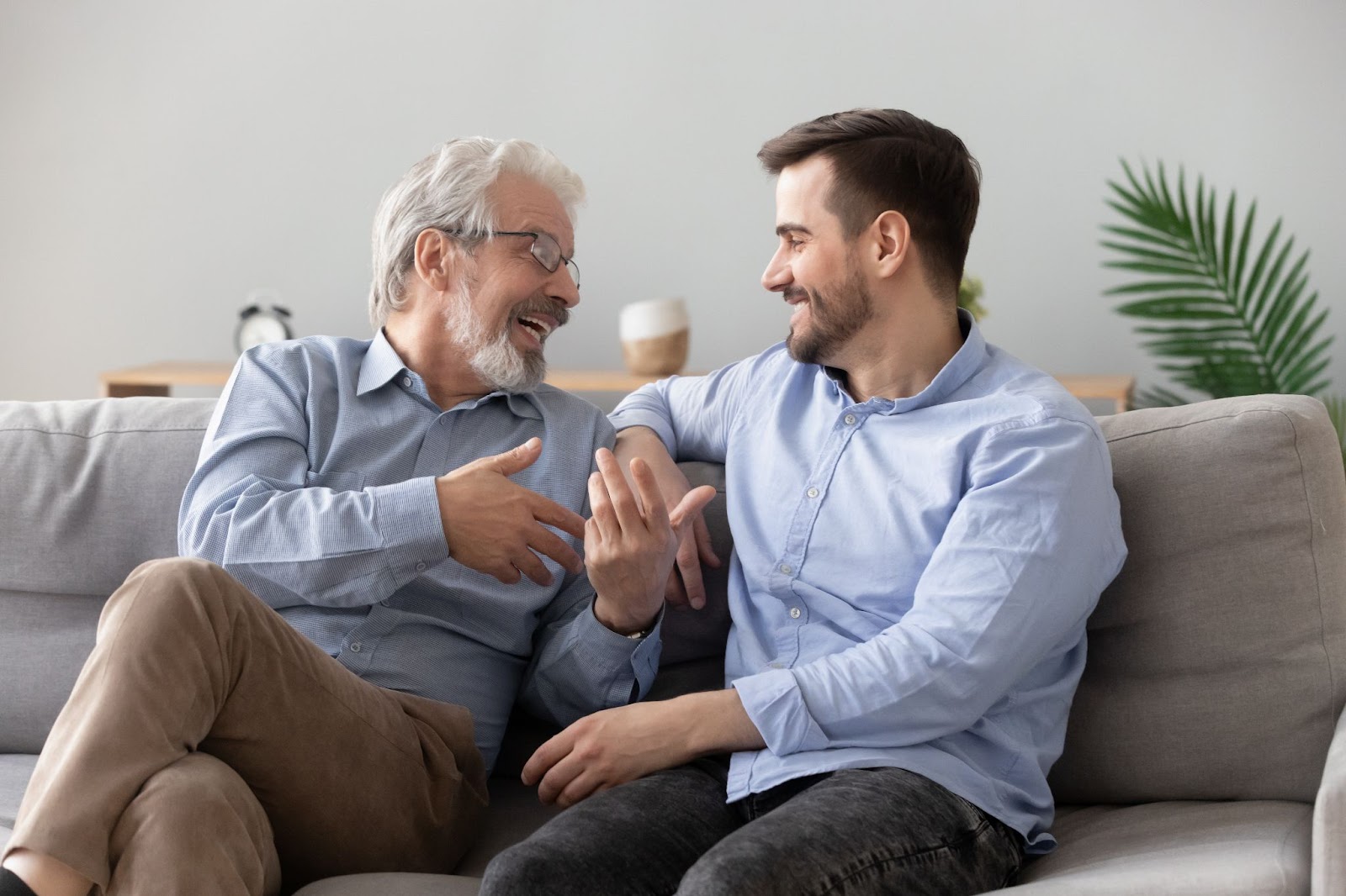 A laughing senior man and his adult son sitting on a couch discussing assisted living.