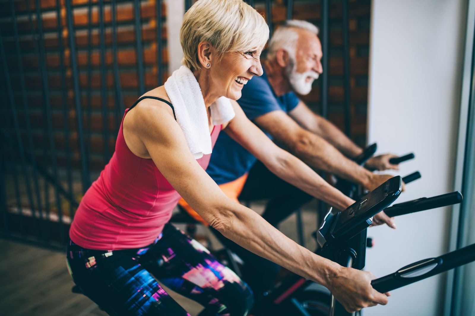 An older woman and man laughing as they exercise together on stationary bikes.