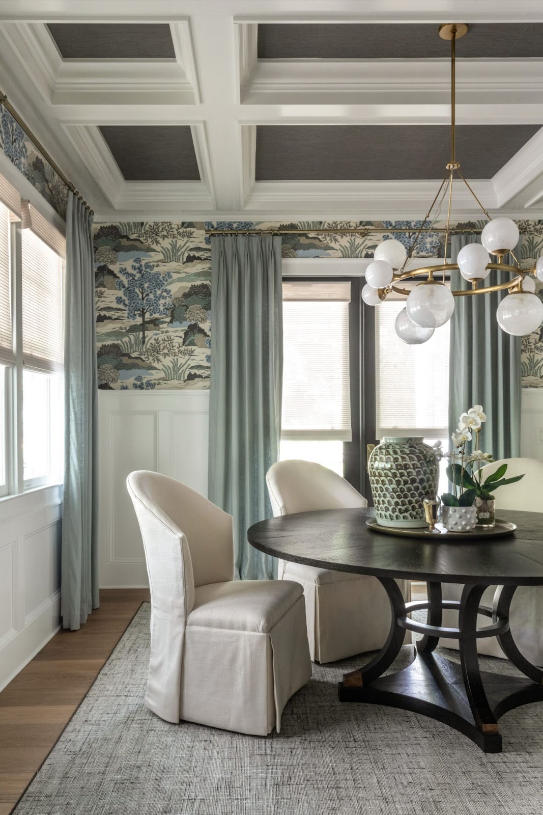 A dining room with a round wooden table, four white upholstered chairs, and a large vase centerpiece. The room has a coffered ceiling, traditional wallpaper with a nature theme, and light blue curtains framing a window.