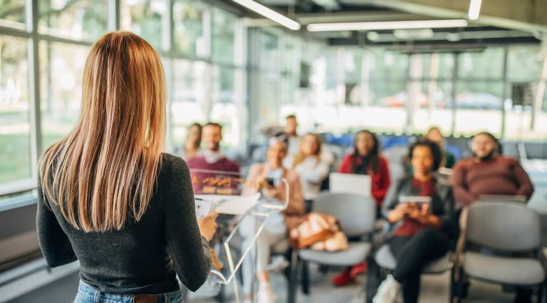 Une femme blonde en pull noir donne une conférence devant un groupe de personnes assises dans une salle de classe moderne avec de grandes fenêtres. Elle se tient derrière un pupitre et utilise des notes pour sa présentation.