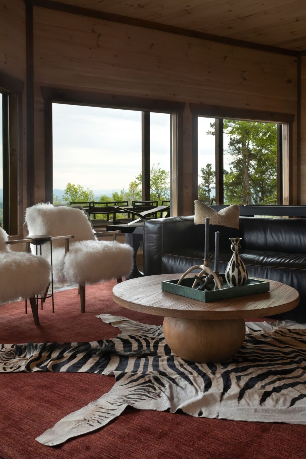 A living room with large windows showcasing a scenic view. The room features a black leather couch, two white fluffy armchairs, a round wooden coffee table, and a zebra print rug. The walls and ceiling are covered in natural wood paneling, contributing to the rustic feel.