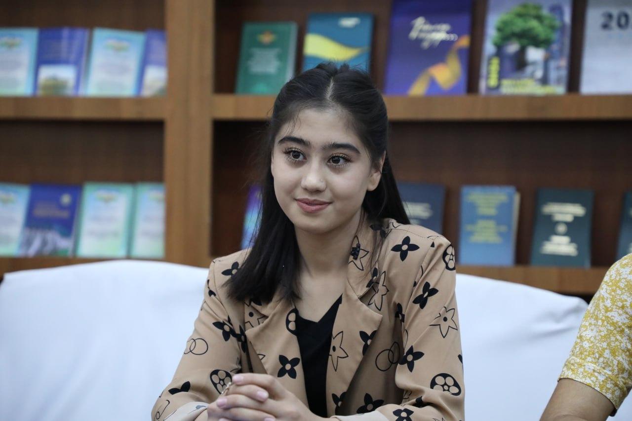 Central Asian woman with long dark straight hair, brown eyes, and a brown suit coat with black star patterns and a black blouse. She's seated on a white couch in front of a wooden bookshelf. 