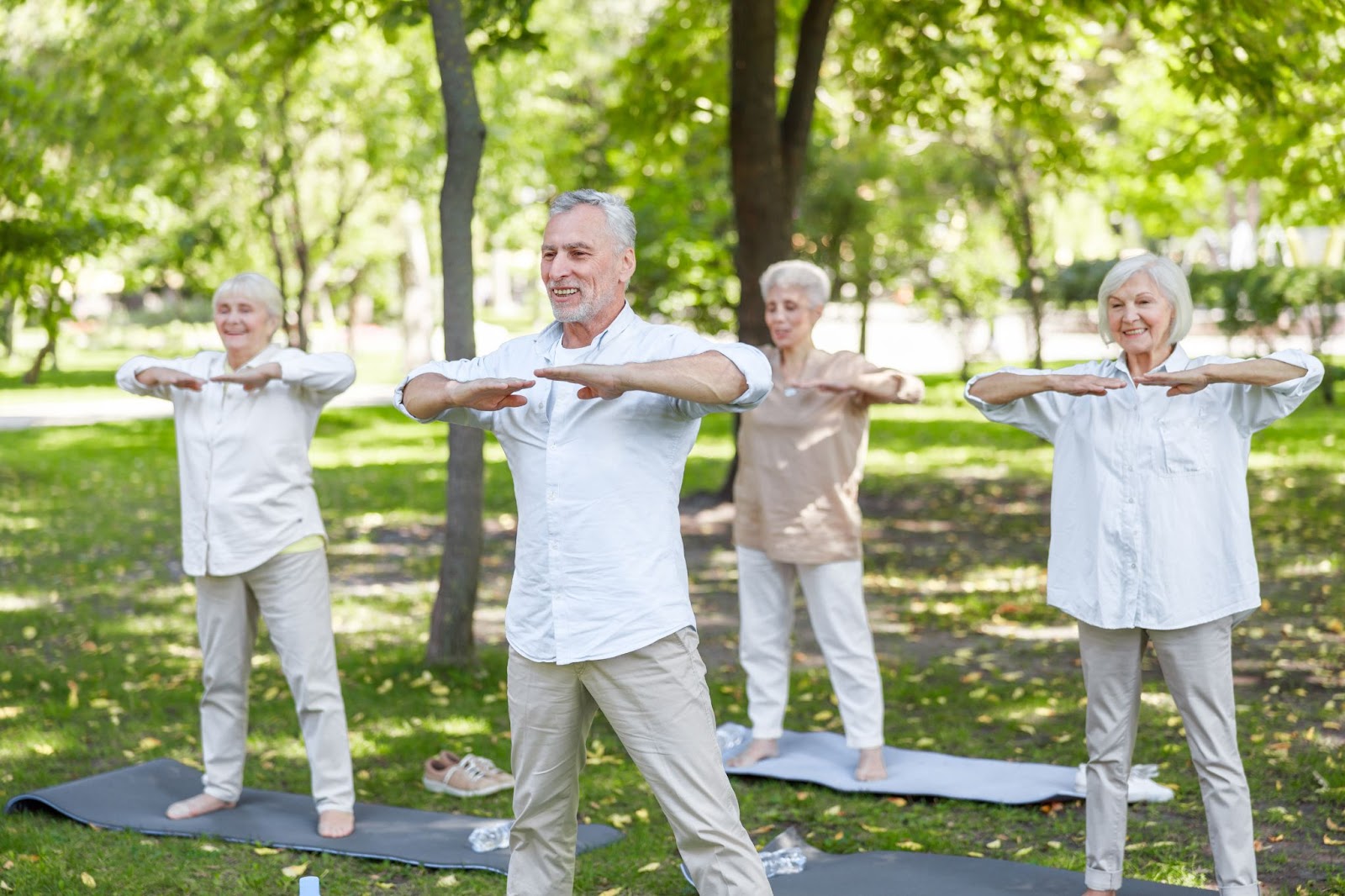 A group of four seniors dressed in white linen practice Tai Chi in an open, green public park. They’re barefoot and smiling.
