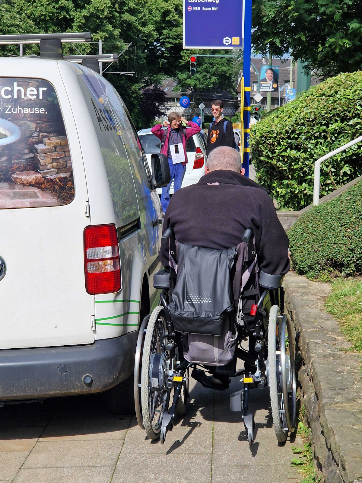 Ein Rollstuhlfahrer versucht zwischen einer Mauer rechts und einem auf dem Gehweg parkenden Transporter links durchzukommen.