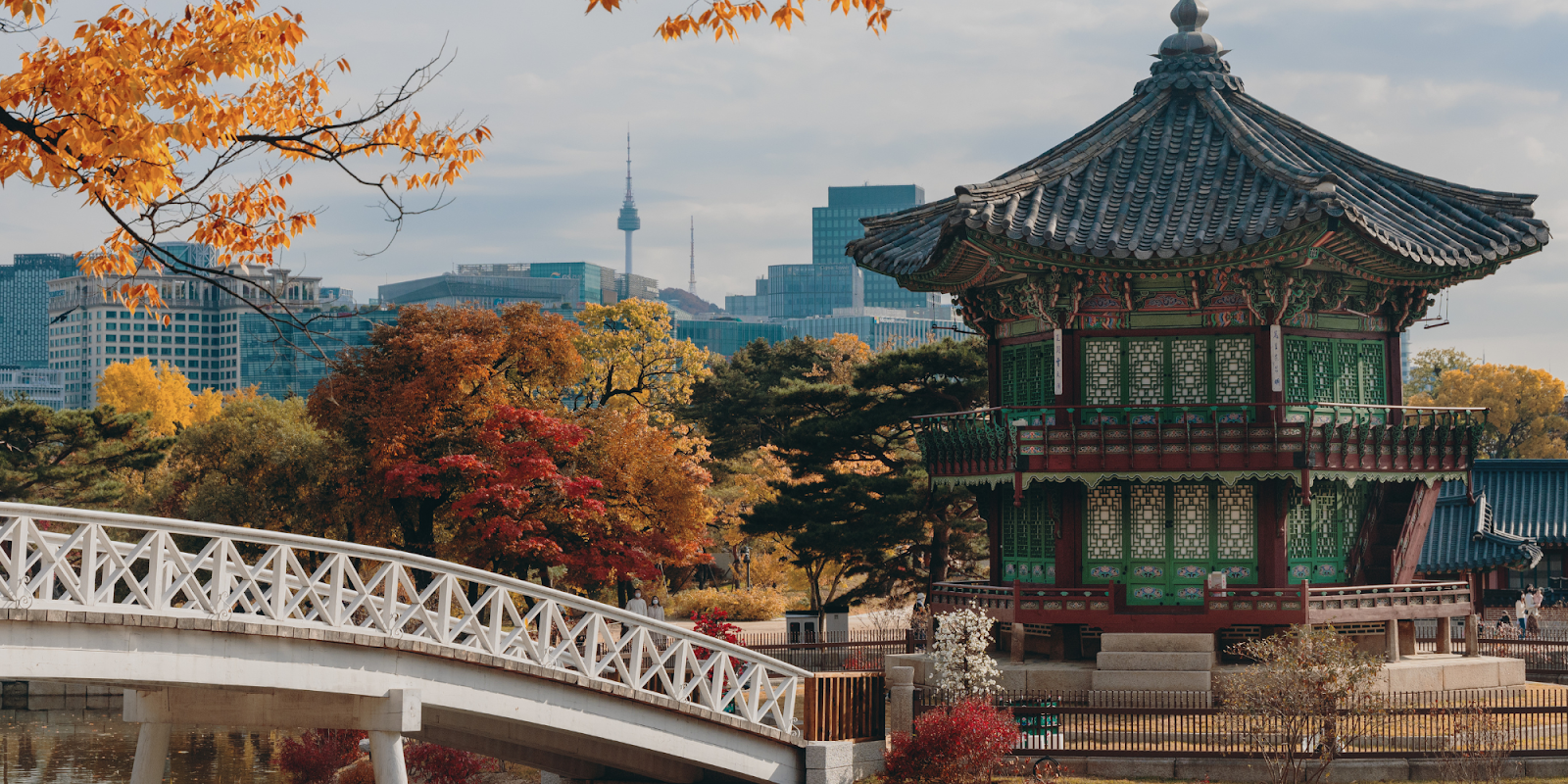 A picturesque autumn scene in Seoul, South Korea, featuring the traditional architecture of a pavilion surrounded by vibrant fall foliage. The pavilion is situated near a serene pond, with a white bridge crossing over. In the background, the modern skyline of Seoul, including the Namsan Seoul Tower, contrasts with the historic and natural elements in the foreground. The colorful leaves and clear sky add to the beauty of this harmonious blend of old and new.