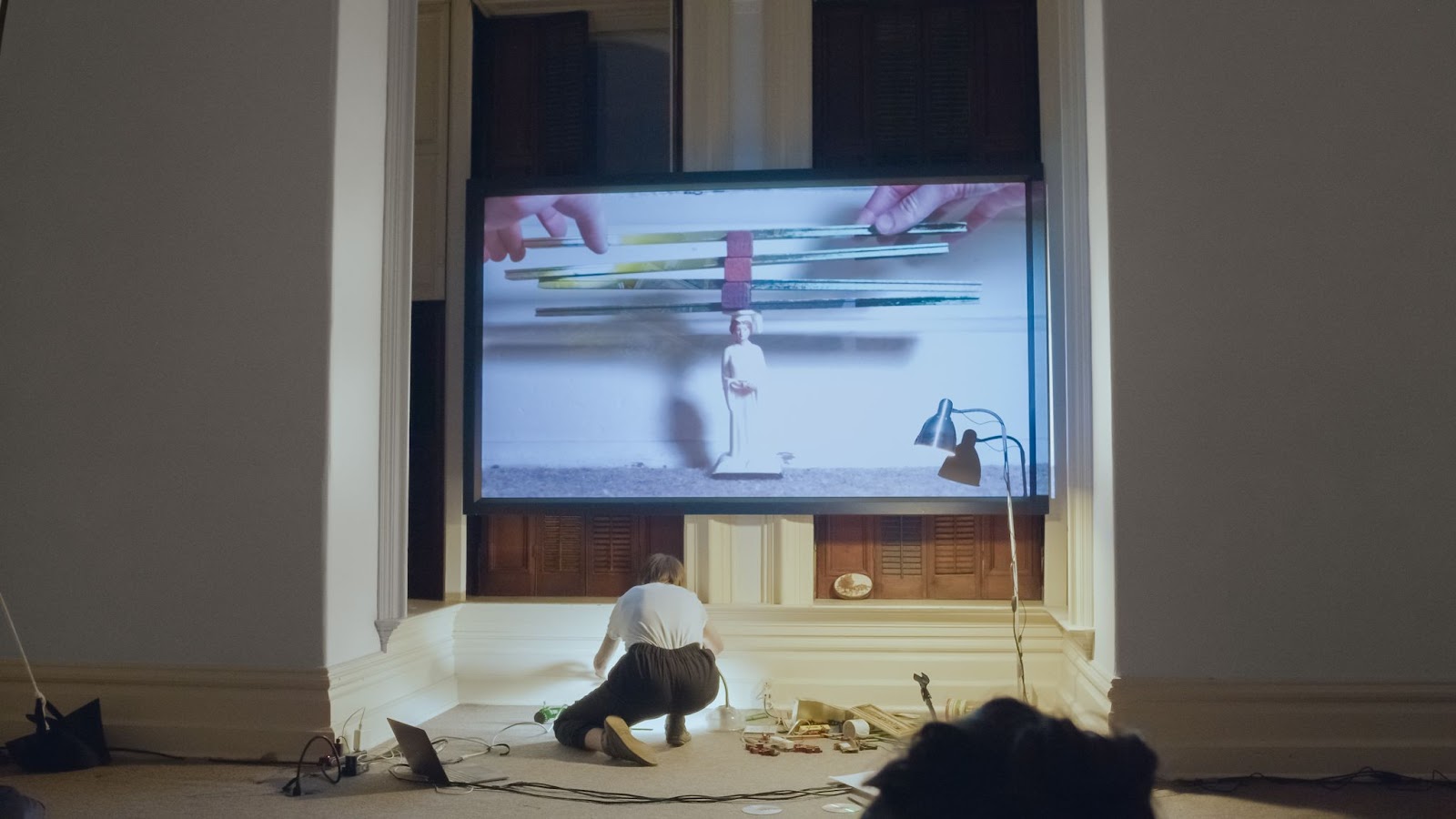 Image: Camille Casmier performs “What Do I Owe Ya” at PS1 Close House. A performer crouches in front of a projector with their back facing the camera. They are arranging small objects underneath light from a desktop and tabletop lamp. There is a small pile of oddities to their right and a MacBook to their left. On-screen, the performer’s hands balance green “Lincoln Logs” atop a plastic figurine of a graduate. Photo courtesy of the artist.