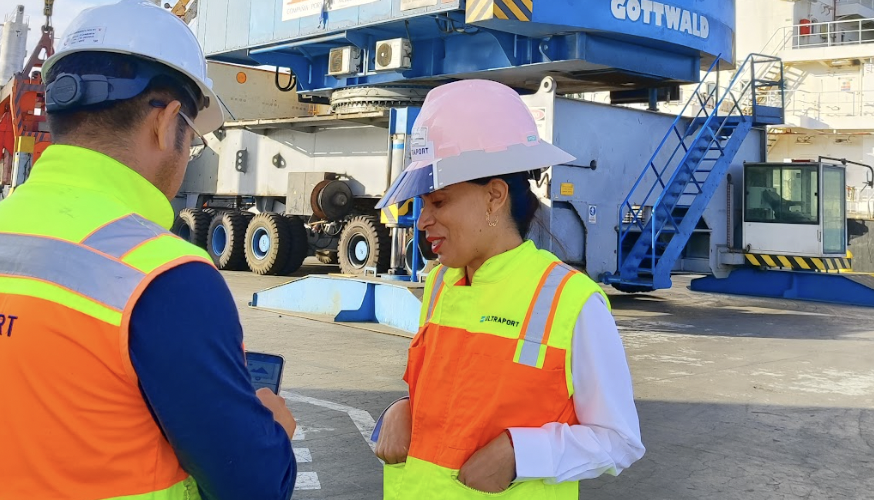 Two people in vests looking at a phone with machinery in the background. 