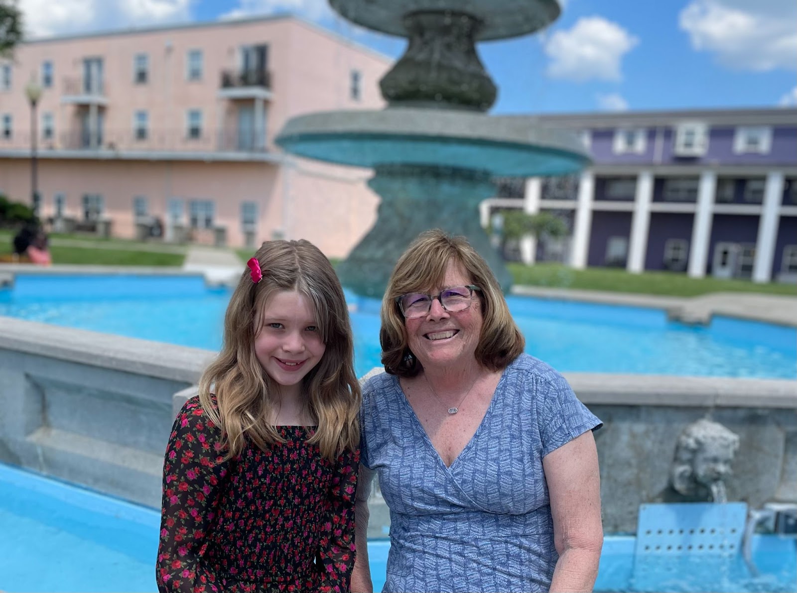 A girl and her grandma sitting next to each other in front of a fountain, smiling. It's a sunny day with some clouds.