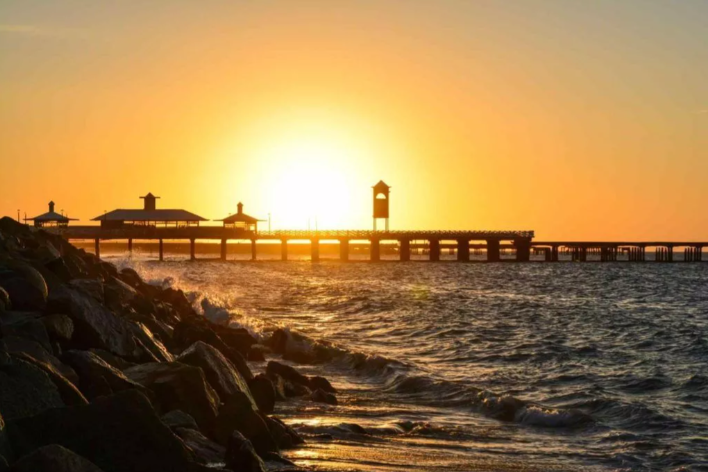 Pôr do sol na Praia de Iracema, Fortaleza, com destaque para a vista lateral da Ponte dos Ingleses. A estrutura se ergue sobre o mar por meio de colunas e está ofuscada pelo brilho do sol.