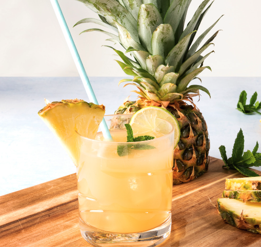 A short glass with a yellow mocktail garnished with pineapple, lime, and a straw, and a whole pineapple in the background