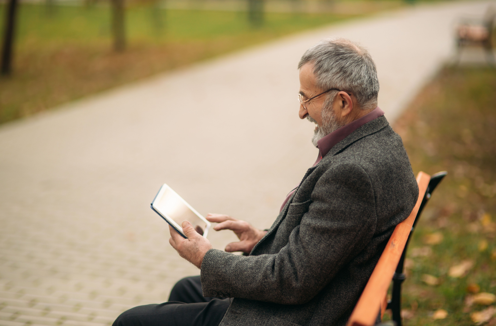 A senior person sits on a park bench with reading glasses smiling down at their e-reader.