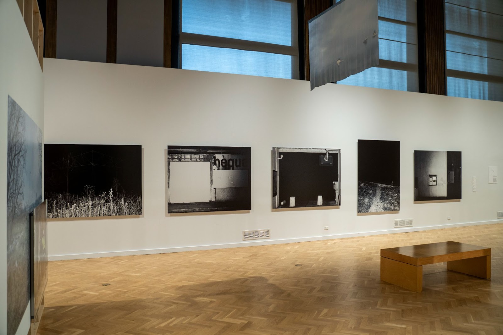 Image: Installation view, Karim Kal, Ligne Dée (2017). Five large black-and-white photographs with images of and around stations on the outskirts of Paris. Above, one of Rebecca Topakian’s silk flags is hung from the ceiling. On the adjacent left wall is a glimpse of Gilberto Guïza-Rojas’ collages. Courtesy of Villa Albertine.