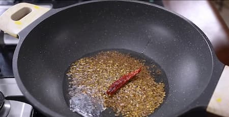 A hot pan with cumin seeds, dry red chili, and crushed coriander seeds being sautéed in oil for the tadka.