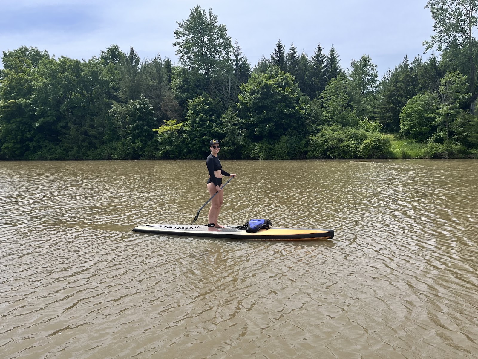 Tarzan paddles on a calm lake or river surrounded by lush green trees and forests. She is wearing a black top and shorts and is standing and using a paddle on a large paddleboard while enjoying the peaceful, natural surroundings.