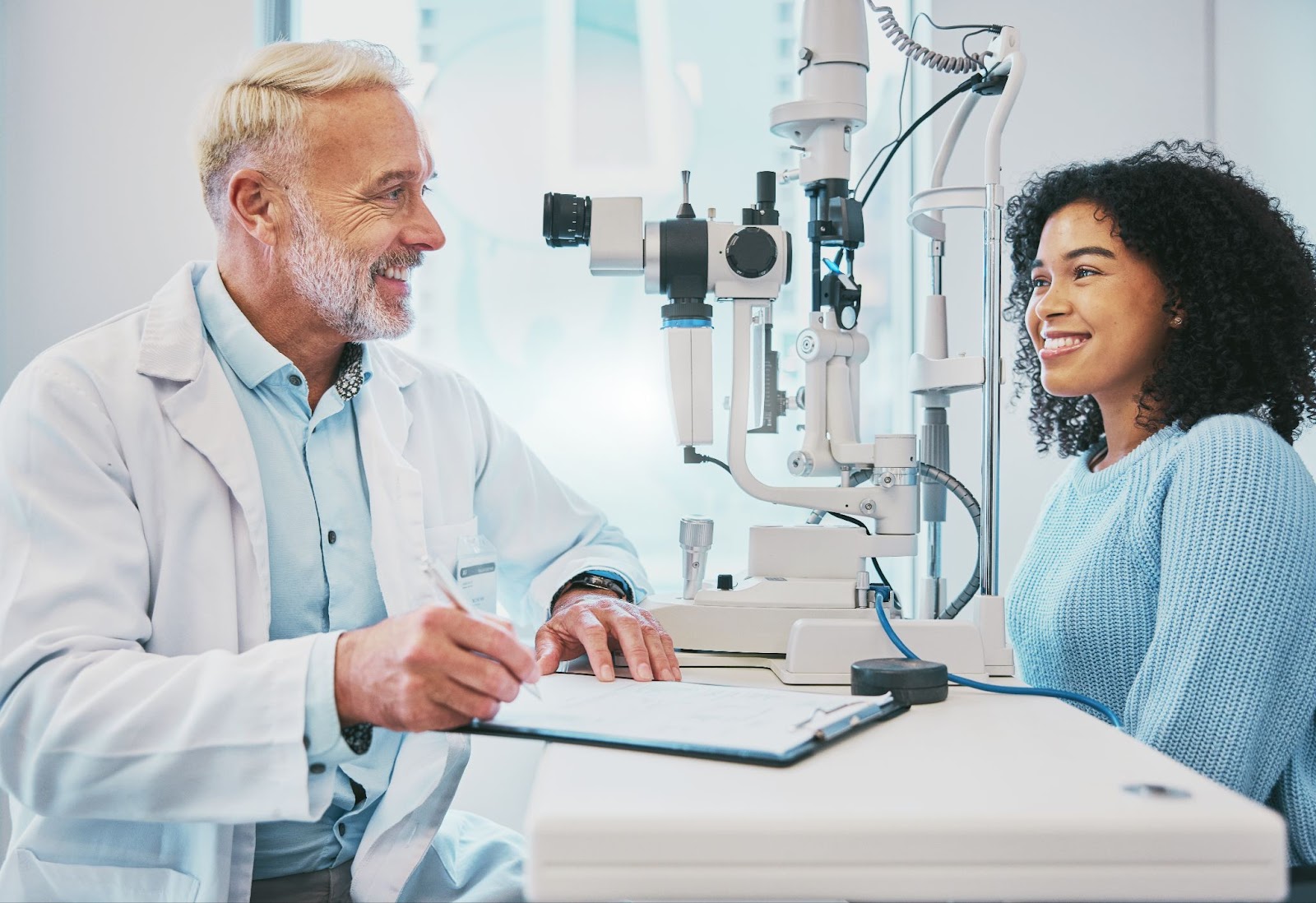 A mature optometrist and a young patient smiling at each other while discussing the results of her eye exam.
