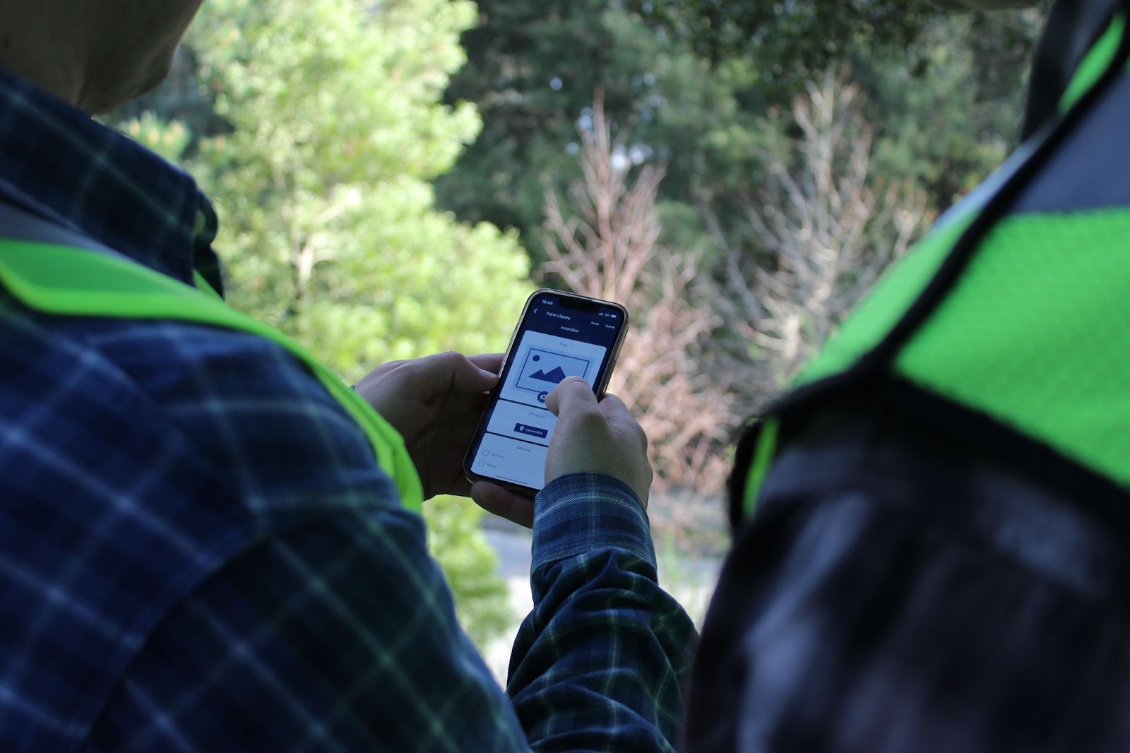  Two men in vests holding and looking at a cell phone with trees in the background. 