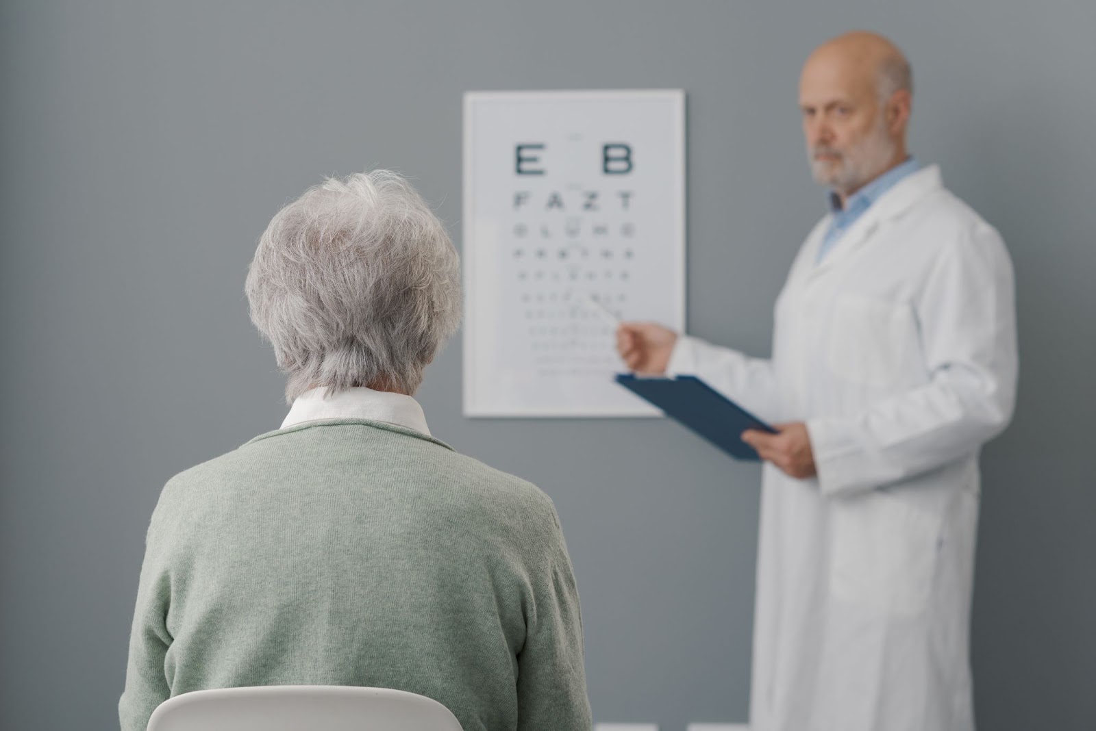 An elderly patient sitting in front of an eye chart while an eye doctor points to it during an eye exam.