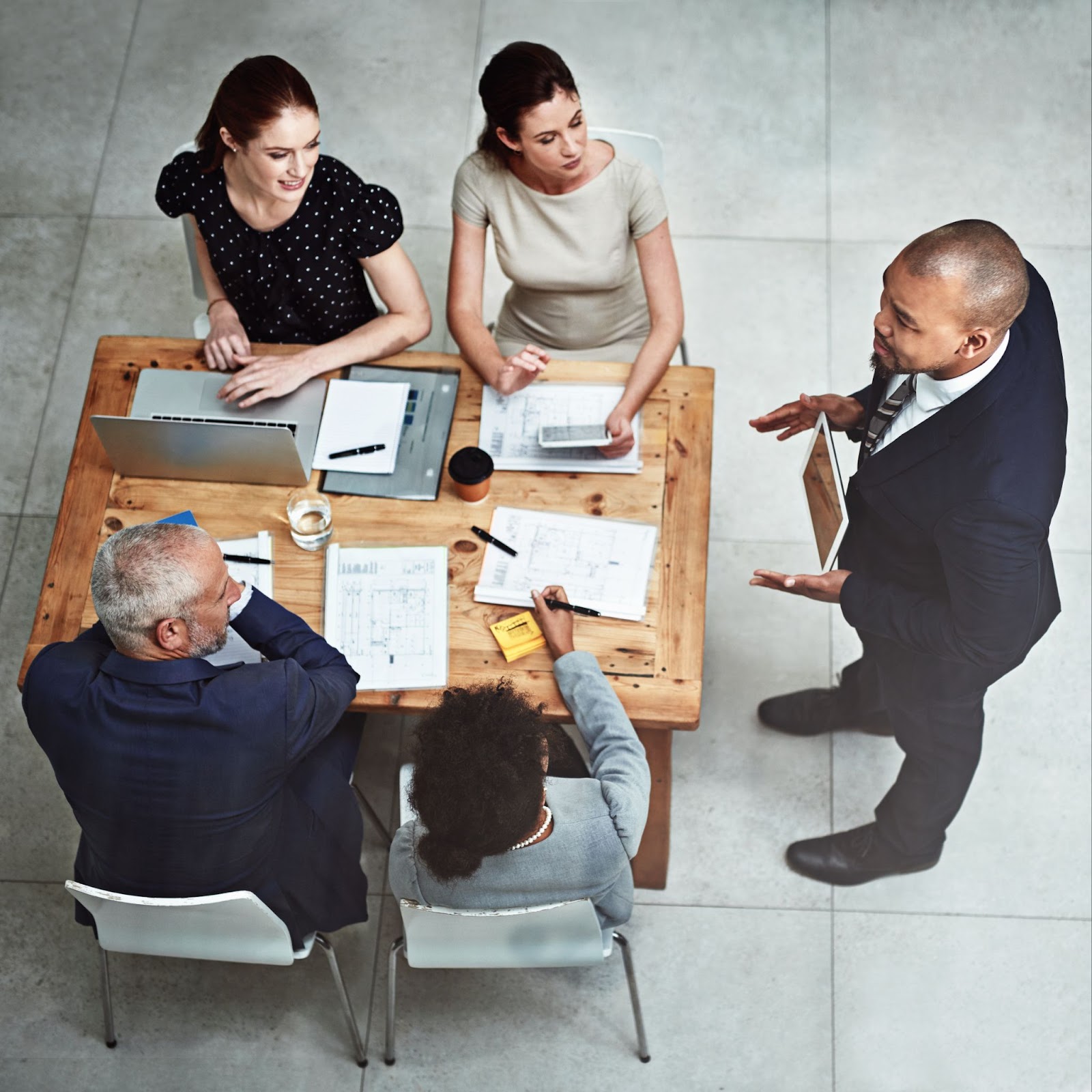 A top shot of a businessman standing in front of four seated colleagues during a meeting, showing them something on a digital tablet. The table is cluttered with a laptop, documents, and pens, emphasizing the collaborative nature of their business formation discussion.