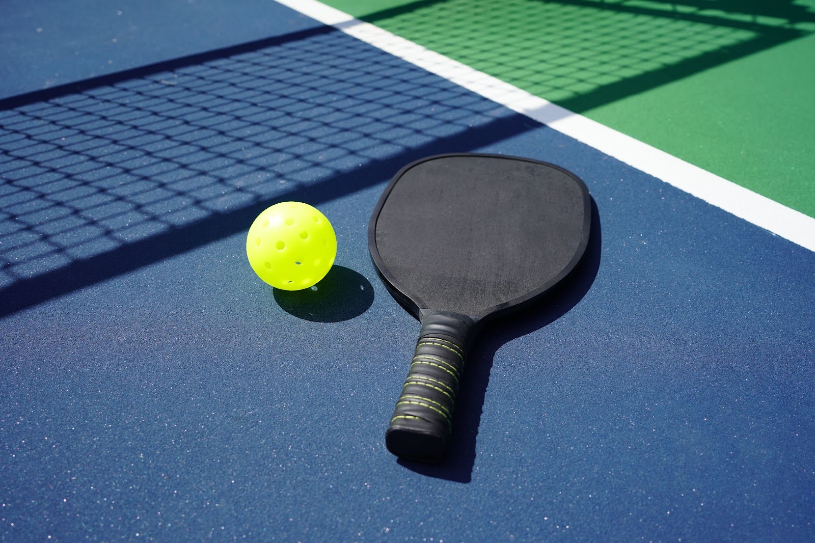 A close-up image of a pickleball paddle and ball lying on the ground in an outdoor court.