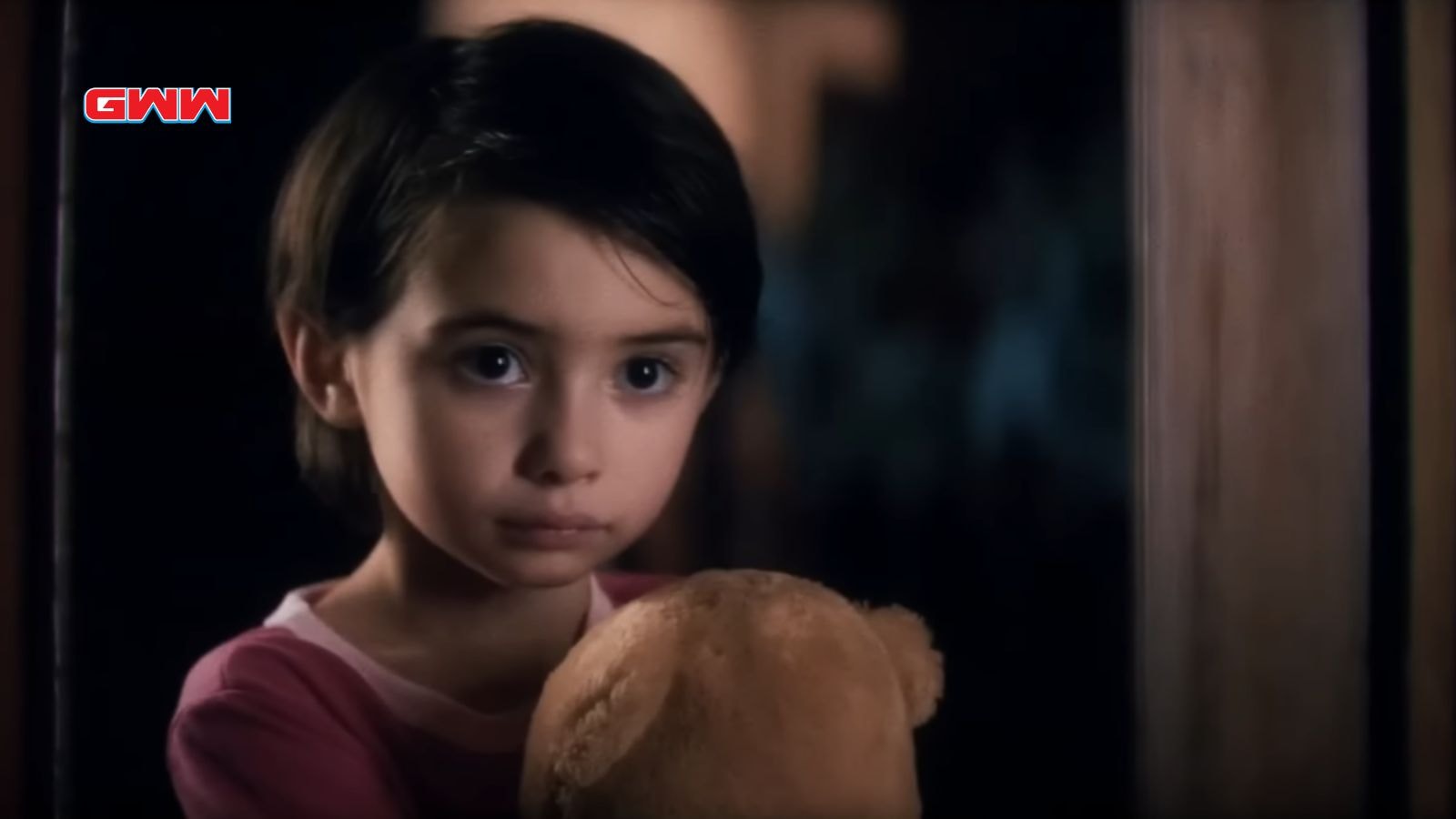 Young girl clutching a teddy bear, looking anxious in dim light.