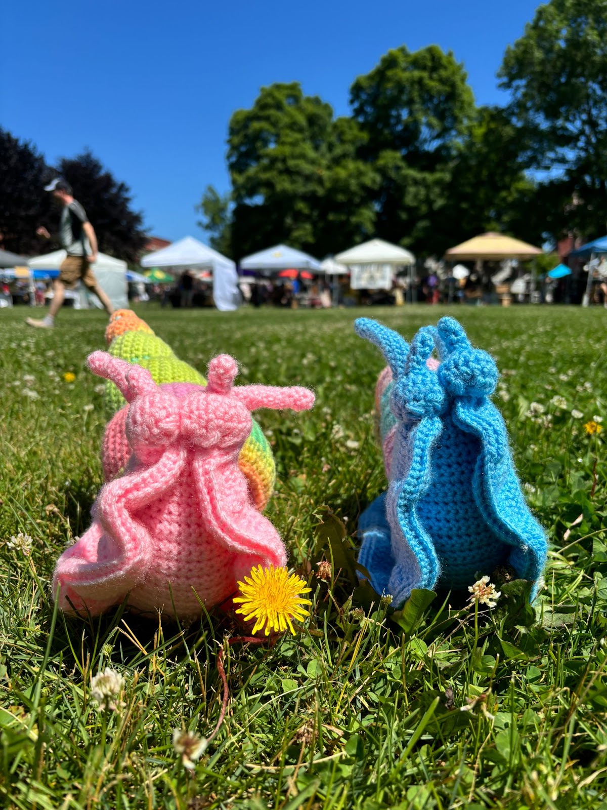 Two crochet snails, sitting on a grass field with a dandelion. There are tents and market stalls behind them. 